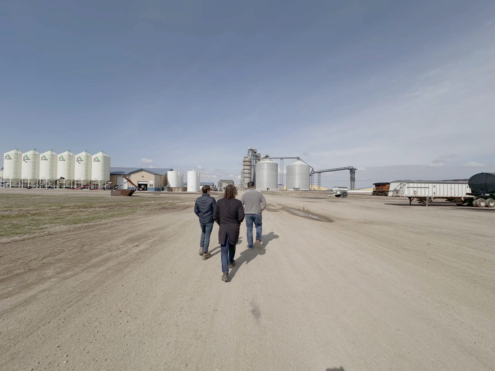 Three people walking across a gravel yard toward a grain elevator